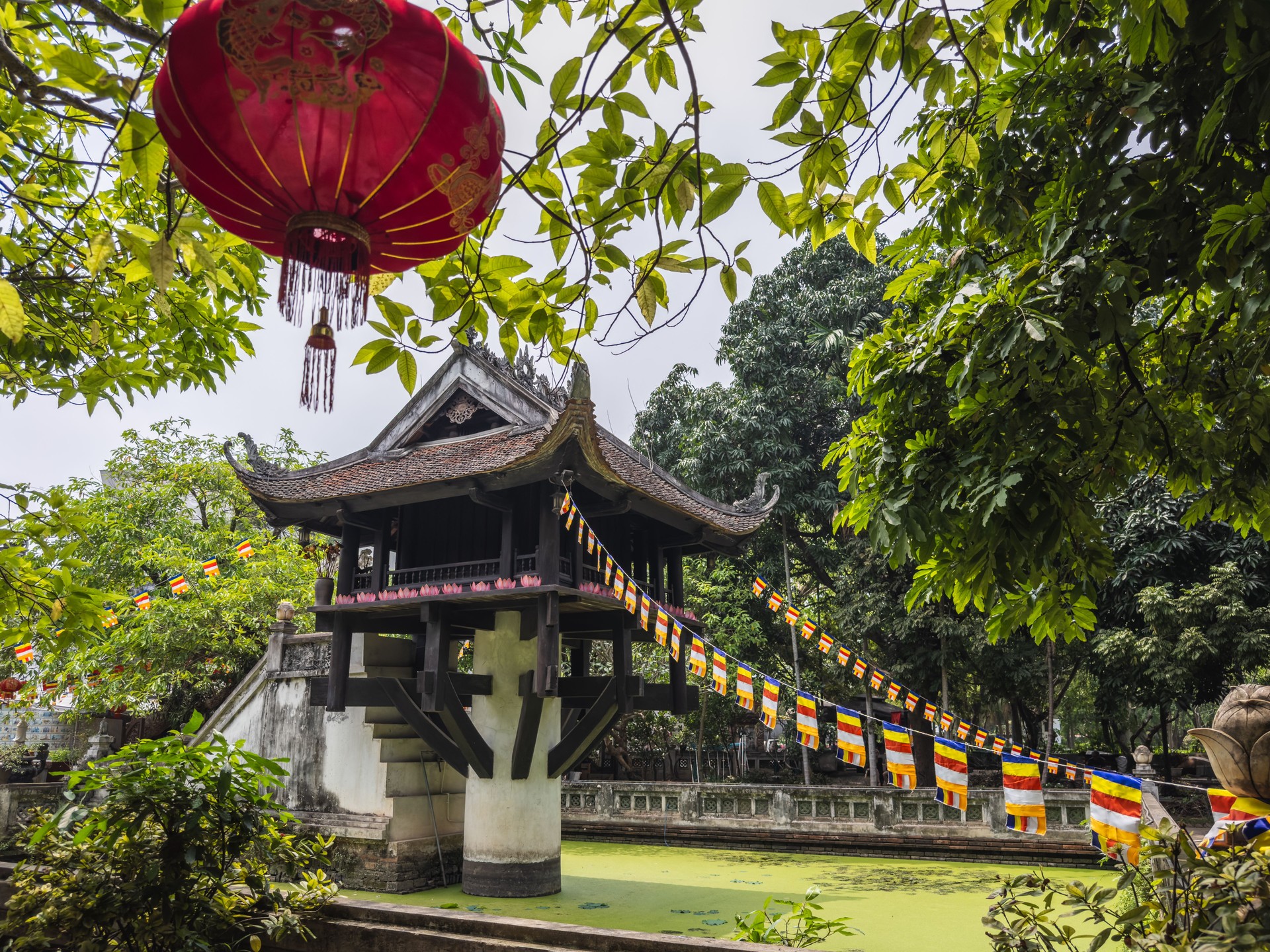 Historic One Pillar Pagoda in Hanoi, Vietnam, uniquely rising from a lotus pond, framed by vibrant green leaves and a traditional red lantern. Colorful Buddhist prayer flags stretch across this iconic