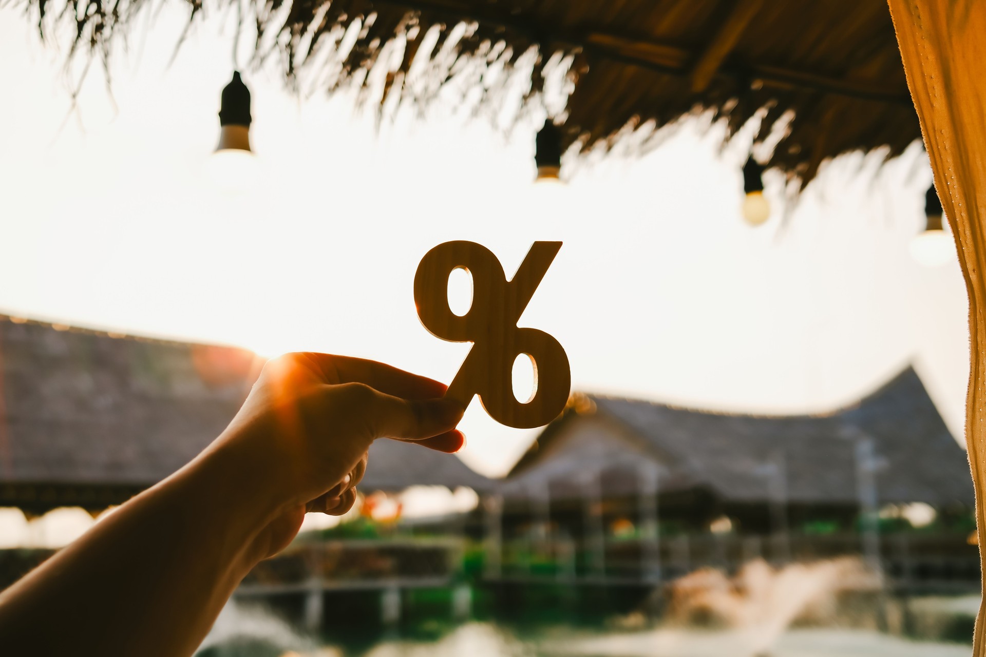 Hand holding a wooden percent sign with tropical resort background at sunset, symbolizing travel discount, hotel promotion, seasonal sale, tourism marketing, and vacation deals in Thailand.
