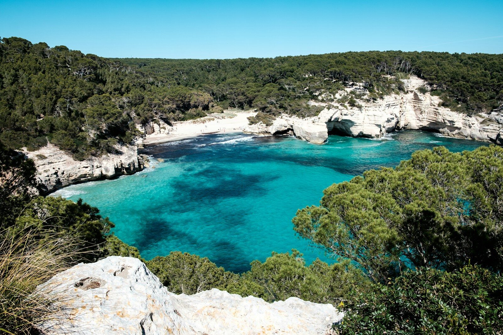 Promotional travel image showing Menorca from Dublin. Travel dates: Wed 6 May 2026 – Wed 13 May 2026. Flights – return (direct). Price €58. Mediterranean beach background with turquoise water, coastal cliffs, Spanish flag, seashells and summer accessories on sand.